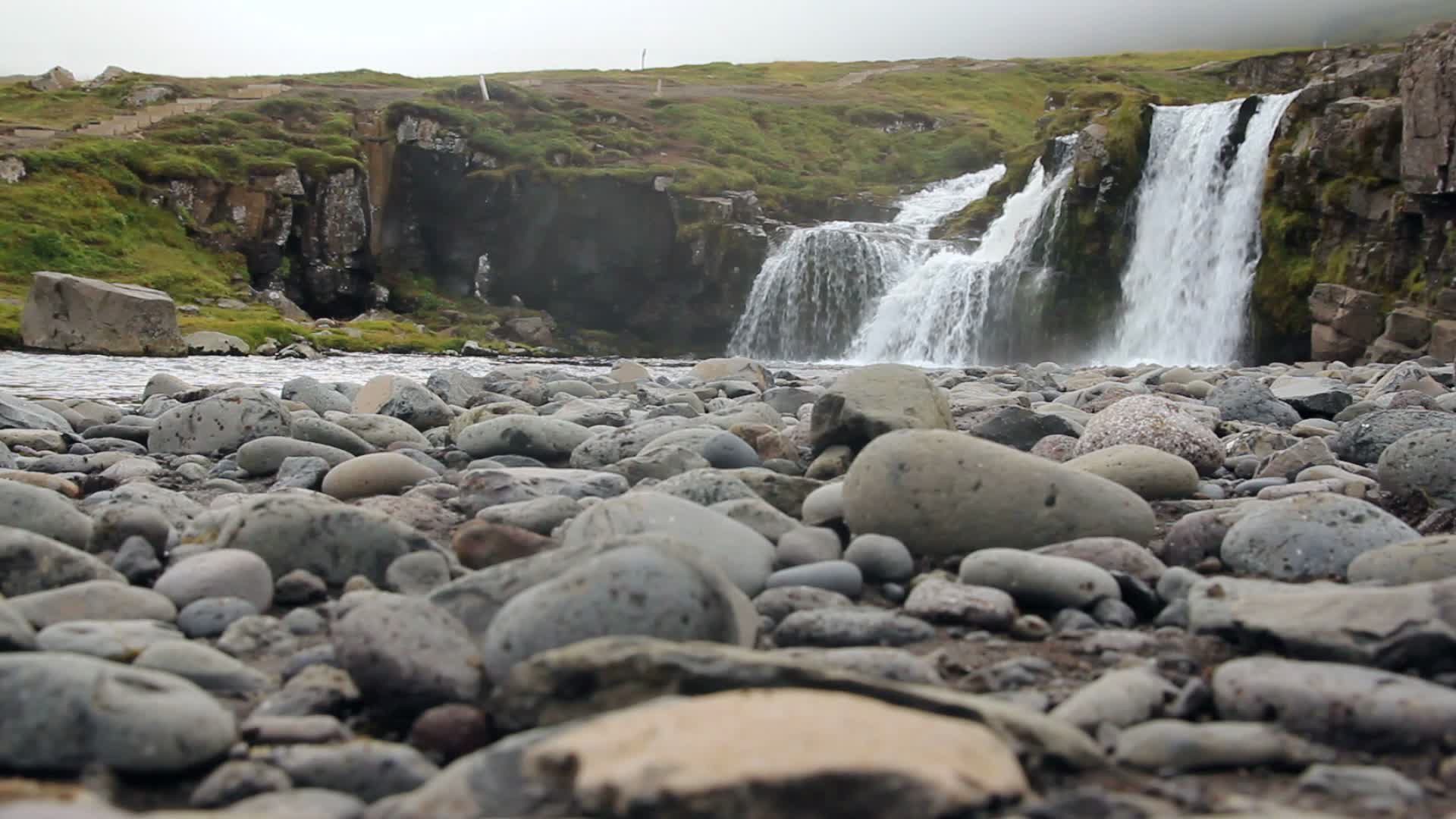 Cascade brumeuse s'écoulant sur des falaises rocheuses sombres, galets gris au premier plan, mousses vertes